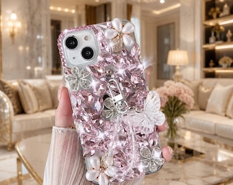 May include: A clear phone case decorated with numerous pink faceted crystals, white pearl-like flower accents, and a translucent butterfly charm. The case features a silver metal ring holder attached to the back, creating a highly reflective surface.