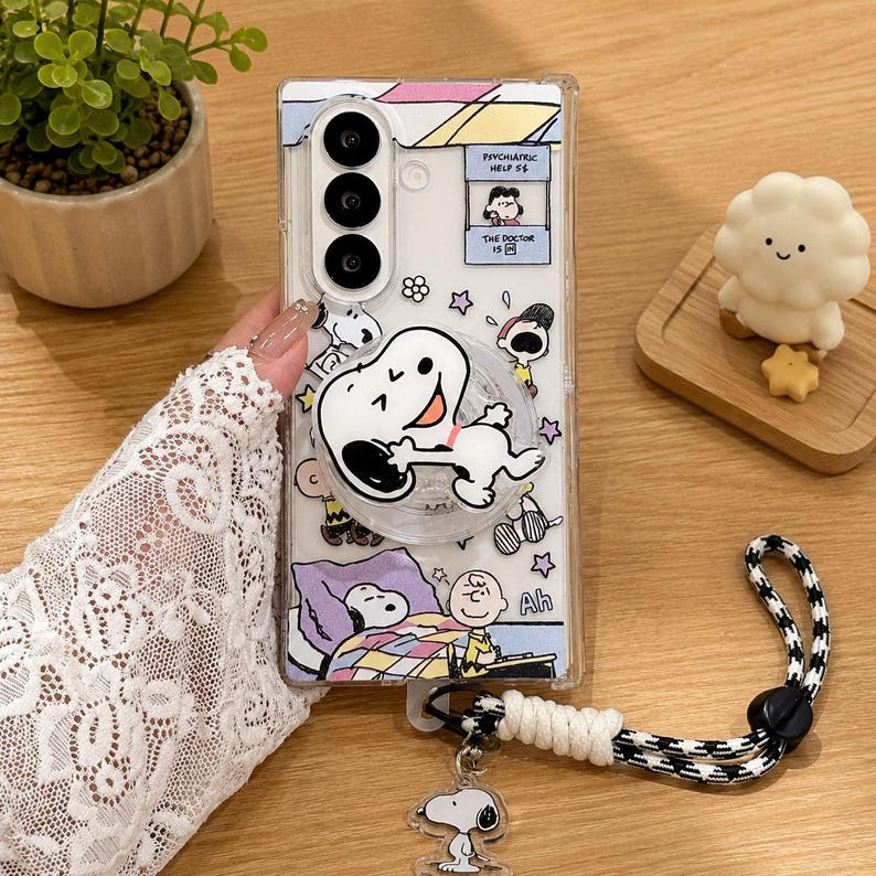 May include: A clear smartphone case featuring Peanuts characters including Snoopy and Charlie Brown. The case includes a circular Snoopy phone grip, a wrist strap with a black and white pattern, and a Snoopy charm. Text reads Psychiatric Help 5 cents.