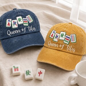 May include: A dark blue denim baseball cap and a mustard yellow baseball cap, both featuring embroidered Mahjong tiles and the white cursive text Queen of Tiles. Three individual white Mahjong tiles are placed in front of the caps.