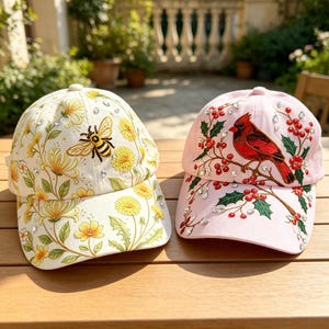 May include: Two baseball caps. The left cap is cream-colored with yellow dandelion flowers, green leaves, and an embroidered bee. The right cap is light pink with an embroidered red cardinal bird, green holly leaves, red berries, and clear rhinestones.
