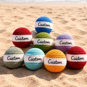 May include: A collection of handmade crochet balls in various colors, including red, blue, green, and yellow, each with a white band featuring the word "Custom". The balls are arranged on a sandy beach.