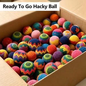 May include: A cardboard box filled with numerous small, round, crocheted hacky balls in a variety of vibrant colors and patterns. The text 'Ready To Go Hacky Ball' is printed at the top of the image. Each ball features intricate yarn stitching.