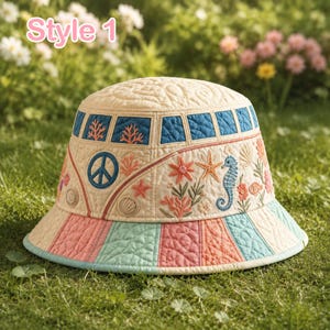 Puede incluir: Un sombrero de pescador acolchado en color crema con el texto Style 1, un símbolo de paz azul y bordados de vida marina que incluyen un caballito de mar, una estrella de mar y coral. El ala tiene rayas verticales alternas en azul claro, rosa y crema.