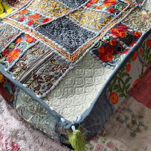 May include: A square floor cushion featuring a patchwork design of various embroidered fabrics in shades of gray, orange, red, and gold. The surface displays intricate floral patterns, geometric stitching, and a small green tassel at the corner.