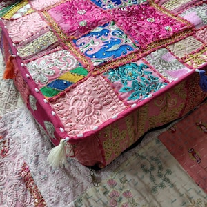 May include: A square floor cushion featuring a patchwork design of various pink, blue, and gold fabrics. The surface is decorated with intricate embroidery, sequins, and metallic thread work. The edges are finished with white beads and tassels.