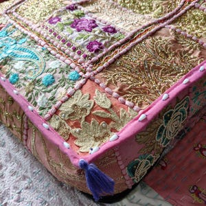 May include: A square floor cushion featuring a patchwork design of pink, peach, and gold fabric panels. The surface is decorated with intricate embroidery, metallic gold thread, sequins, and small white beads, finished with a purple corner tassel.