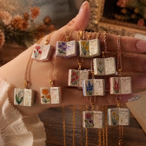 Op de afbeelding: Een collectie goudkleurige kettingen met miniatuur open boekhangers. Elke hanger toont een handgeschilderde bloem en de naam van een maand, waaronder januari, februari, maart, april, mei, juni, juli, augustus, september en november.