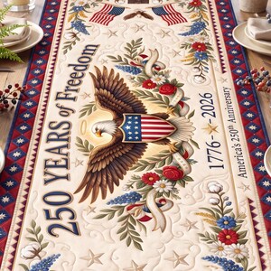 May include: A cream-colored table runner with a patriotic design. It features an American eagle, flags, and floral elements. Text reads "250 Years of Freedom" and "1776-2026". The runner is bordered with a red and blue pattern.