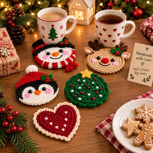 May include: A collection of five round and heart-shaped crochet coasters featuring holiday designs including a snowman, a reindeer, a penguin, a Christmas tree, and a red heart with white accents, displayed alongside two mugs and cookies.