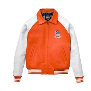 May include: A bright orange leather jacket with white sleeves, a fold-down collar, and a front zipper closure. The chest features a patch with the text AVIREX and a shield graphic. The waist and cuffs have orange and white striped ribbed trim.
