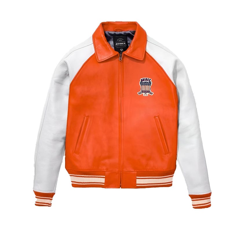 May include: A bright orange leather jacket with white sleeves, a fold-down collar, and a front zipper closure. The chest features a patch with the text AVIREX and a shield graphic. The waist and cuffs have orange and white striped ribbed trim.