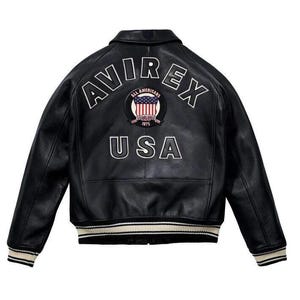 May include: A black leather varsity jacket featuring the white embroidered text AVIREX and USA on the back. A circular patch in the centre displays the American flag with the text ALL AMERICANS ESTABLISHED 1975. The jacket has striped ribbed cuffs.