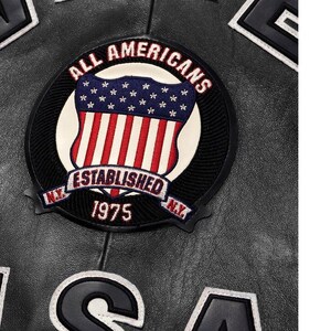May include: A black leather jacket featuring a circular embroidered patch with the text ALL AMERICANS ESTABLISHED 1975 N.Y. and a shield graphic displaying the American flag. Large black letters with white trim are visible at the top and bottom edges.