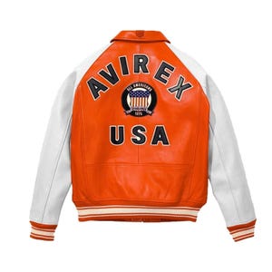 May include: An orange and white leather varsity jacket featuring the text AVIREX and USA in black lettering on the back. A circular patch in the center displays the text ALL AMERICANS ESTABLISHED 1975 with a graphic of the American flag.