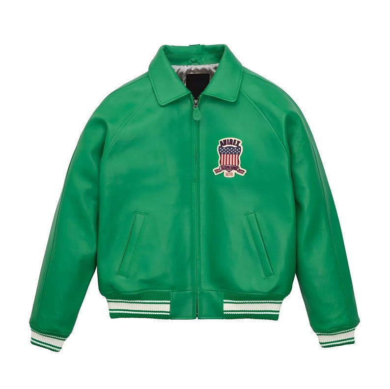 Pode incluir: Um casaco verde brilhante com fecho de correr frontal, gola pontiaguda e punhos e bainha canelados com riscas brancas. No peito esquerdo, existe um emblema com o texto Avirex e um design de escudo com a bandeira americana.