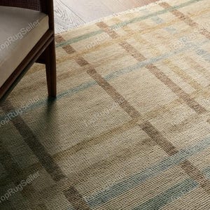 May include: A woven area rug featuring a textured, grid-like pattern in shades of beige, tan, and muted blue. The rug has a visible fringe along the edge and a thick, knotted pile construction. A portion of a wooden chair frame is visible in the corner.