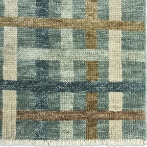May include: A woven rug featuring a grid pattern of intersecting lines in shades of teal, dark blue, light beige, and brown. The textile has a textured, tufted surface and a short fringe along the bottom edge, creating a geometric plaid design.