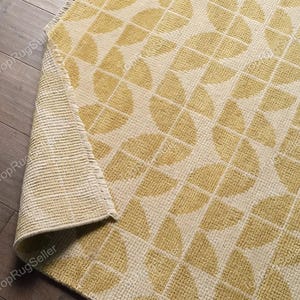 May include: A rectangular area rug featuring a repeating geometric pattern of yellow semicircles on a cream background. The rug has a textured, woven surface and a folded corner revealing a light-colored, grid-like backing with a fringed edge.