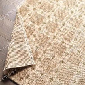 May include: A beige area rug with a repeating grid pattern of squares and small green dots at each intersection. The rug features a textured, tufted pile surface and a folded corner revealing the woven backing and short fringe along the edge.