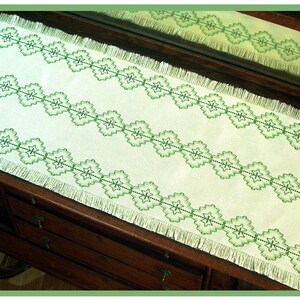 May include: A white table runner with a green embroidered design of repeating diamond shapes and flowers. The runner has a fringed edge.