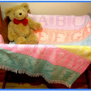 May include: A multi-colored baby blanket with a fringe border, featuring the alphabet in a woven pattern. A brown teddy bear with a red bow tie is sitting on the blanket.