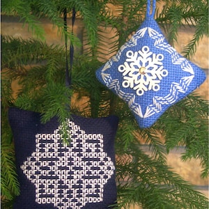 May include: Two blue and white embroidered snowflake ornaments hanging on a green pine tree branch. The ornaments are square shaped and have a white snowflake design in the center.