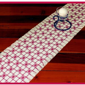 May include: A white table runner with a red diamond pattern and fringe edges. The runner is on a wooden table with a candle holder in the center.