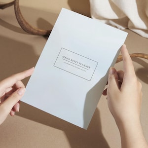 Könnte beinhalten: Ein hellblau-grauer Planer mit dem Titel "Mama Reset Planner" und dem Text "Minimalistischer Self-Care & Wellness Planer". Der Planer wird von zwei Händen vor einem neutralen Hintergrund mit einem dekorativen Element gehalten.