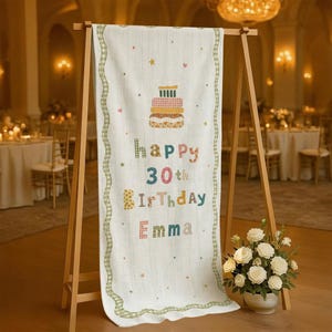 May include: A vertical white fabric banner displayed on a wooden easel, featuring a multi-colored graphic of a tiered cake at the top. The text reads "happy 30th birthday Emma" in various patterned fonts, surrounded by small stars and dots, with a green gingham border.