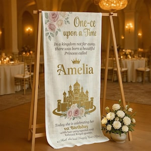 May include: A cream-colored banner with gold text and a castle illustration, displayed on a wooden easel. The banner reads "One-ce upon a Time" and "Amelia", celebrating a 1st birthday. Floral accents and the phrase "Happily Ever After" complete the design.