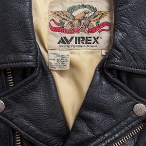 May include: A close-up of a black leather jacket with a cream lining. The jacket displays the "AVIREX" brand label with an eagle emblem. The jacket has a zipper and metal snap details. The label states "100% Genuine Leather".
