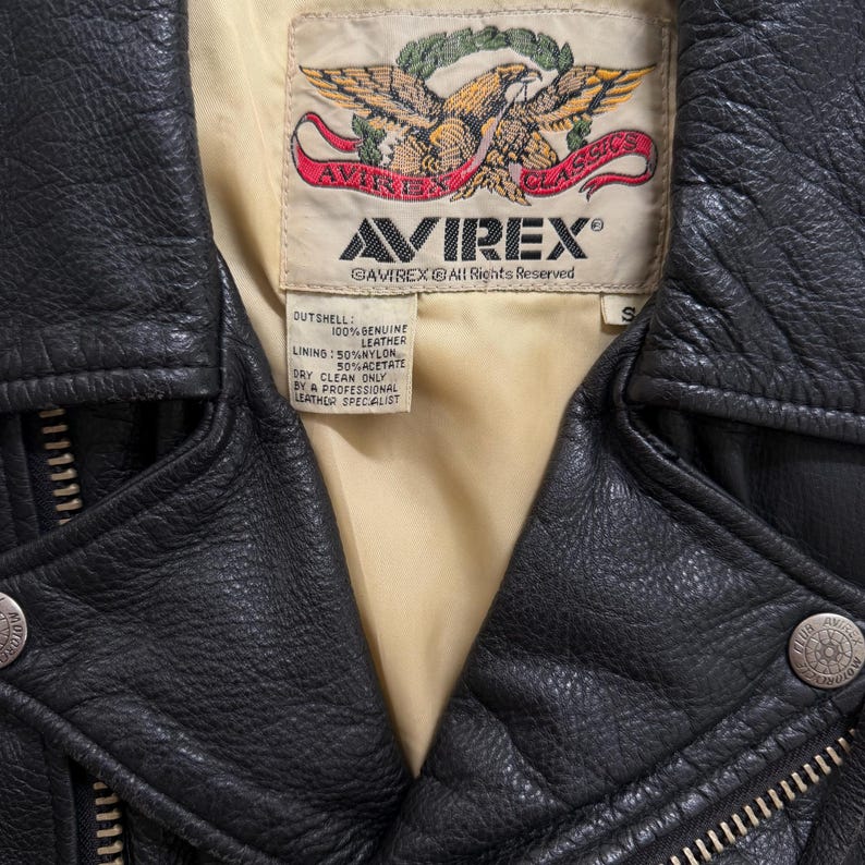 May include: A close-up of a black leather jacket with a cream lining. The jacket displays the "AVIREX" brand label with an eagle emblem. The jacket has a zipper and metal snap details. The label states "100% Genuine Leather".