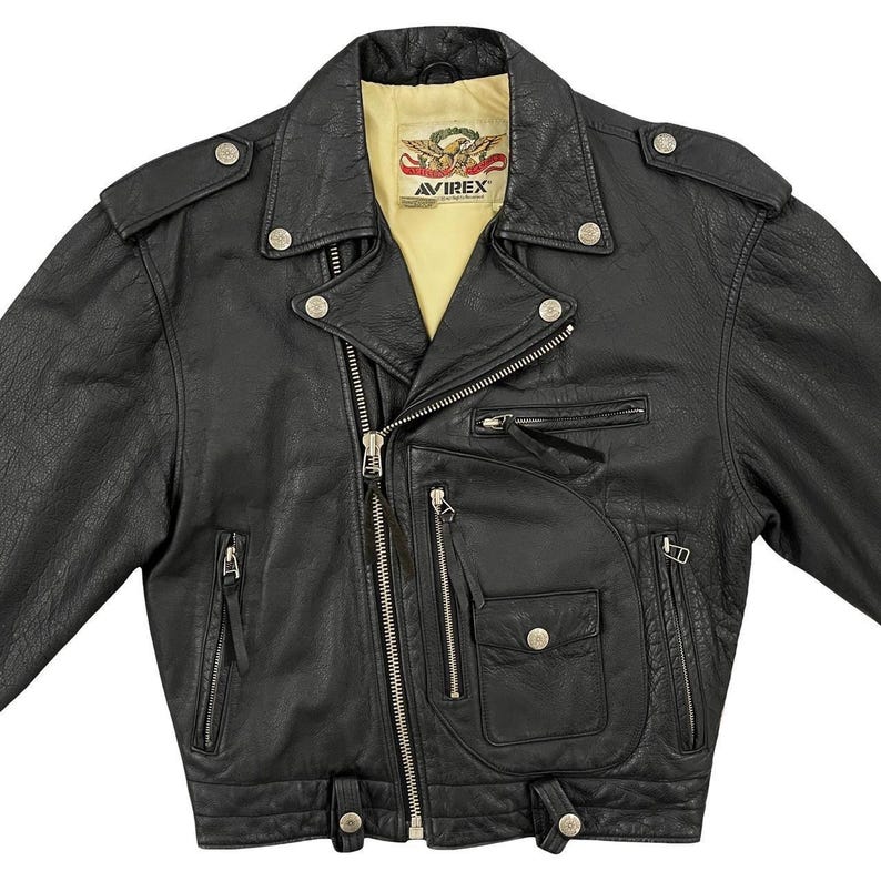 May include: Black leather motorcycle jacket with multiple zipped pockets, snap fastenings, and a notched collar. The jacket features the brand name "AVIREX" on a patch inside the collar. The jacket has a classic biker style.