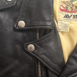 May include: Close-up of a black leather jacket with silver metal buttons and a zipper. The jacket has a textured surface and a yellow lining. The label reads "AVIREX" and "100% Genuine Leather".
