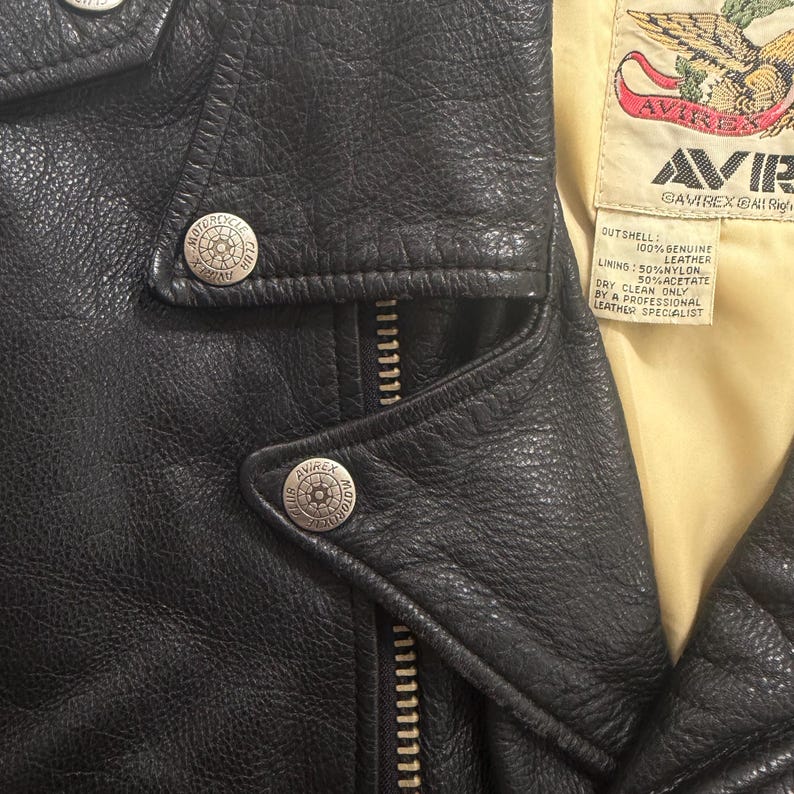 May include: Close-up of a black leather jacket with silver metal buttons and a zipper. The jacket has a textured surface and a yellow lining. The label reads "AVIREX" and "100% Genuine Leather".