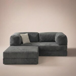 May include: A dark gray corduroy sofa with a matching rectangular ottoman positioned in front. The sofa features two large seat cushions, two back cushions, and thick armrests. A single light beige square throw pillow rests on the left side.