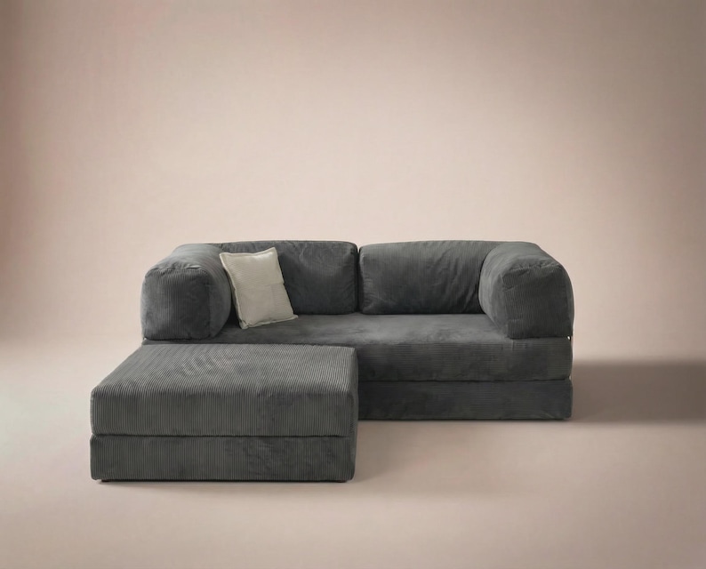 May include: A dark gray corduroy sofa with a matching rectangular ottoman positioned in front. The sofa features two large seat cushions, two back cushions, and thick armrests. A single light beige square throw pillow rests on the left side.