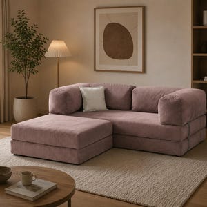 May include: A modular sofa in a dusty rose fabric with a matching ottoman, featuring a low-profile design, thick cushioned armrests, and a single square white throw pillow placed against the backrest, creating a cohesive seating arrangement.
