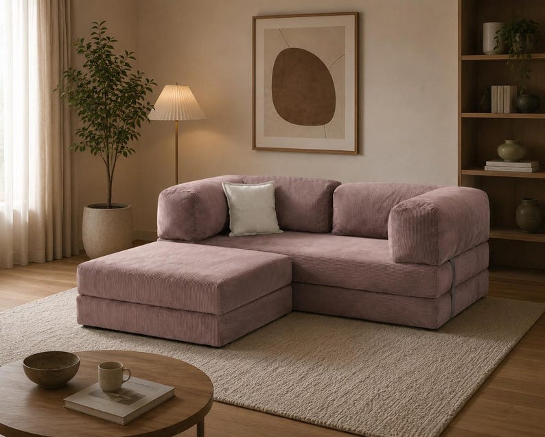 May include: A modular sofa in a dusty rose fabric with a matching ottoman, featuring a low-profile design, thick cushioned armrests, and a single square white throw pillow placed against the backrest, creating a cohesive seating arrangement.