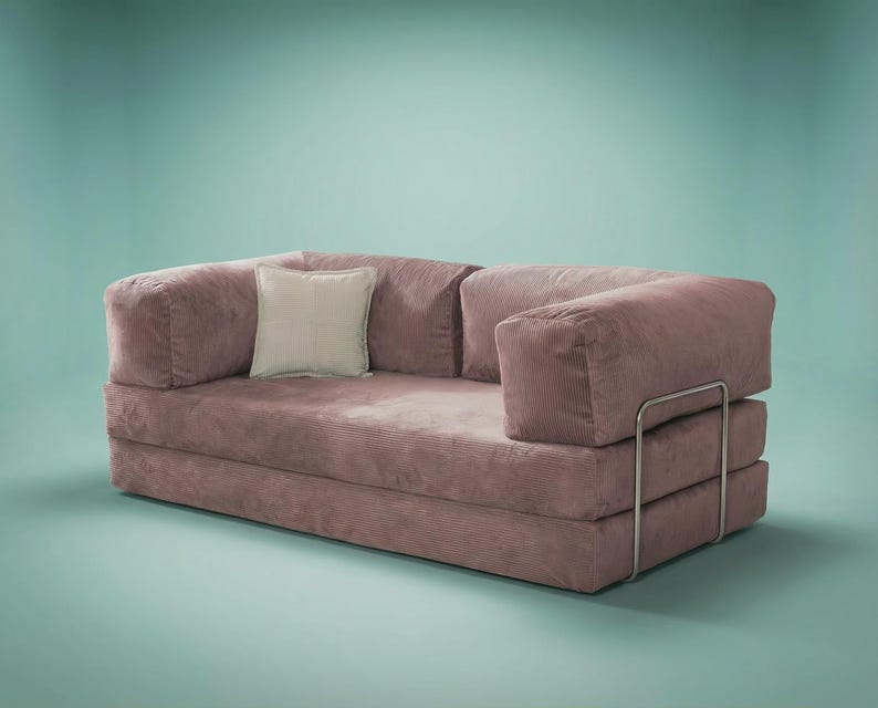 May include: A mauve corduroy sofa featuring a low-profile design with thick, rectangular armrests and a matching backrest. A single square off-white throw pillow rests on the seat. The sofa is supported by a thin, silver-toned metal frame.