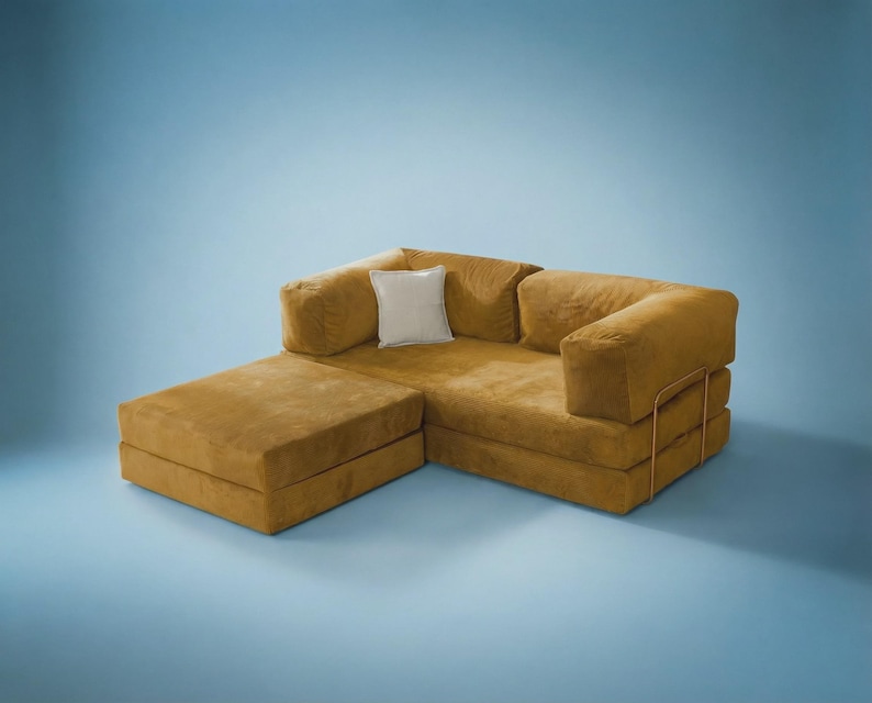 May include: A mustard yellow modular sofa with a low-profile design, featuring a rectangular chaise section, thick cushioned backrests, and a single square off-white throw pillow. The sofa is supported by a thin, gold-toned metal frame structure.