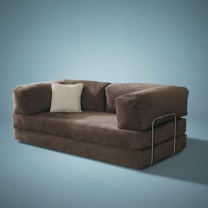 May include: A brown corduroy sofa bed featuring a low-profile design with thick, plush cushions and a single cream-colored square throw pillow. The frame is supported by a visible, thin, silver-toned metal structure on the right side of the base.