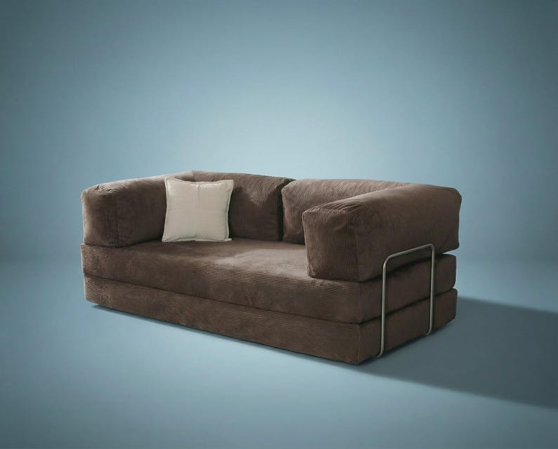 May include: A brown corduroy sofa bed featuring a low-profile design with thick, plush cushions and a single cream-colored square throw pillow. The frame is supported by a visible, thin, silver-toned metal structure on the right side of the base.