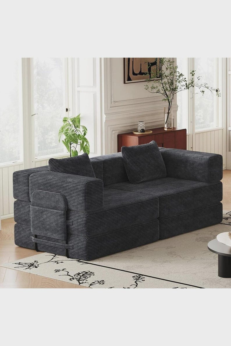 May include: A dark gray corduroy sofa featuring a modular design with thick, rectangular armrests and two matching square throw pillows. The fabric has a distinct vertical ribbed texture, and the structure consists of two main seating sections.