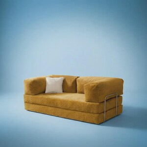May include: A mustard yellow corduroy sofa featuring a low-profile design with thick, rectangular cushions and a single white square throw pillow. The piece is supported by a thin, silver-toned metal frame that wraps around the exterior sides.