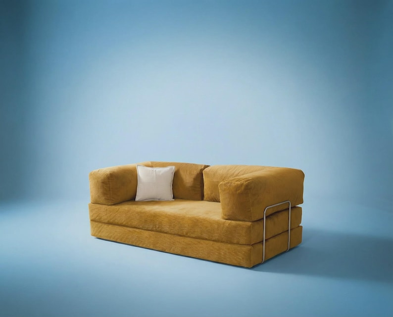 May include: A mustard yellow corduroy sofa featuring a low-profile design with thick, rectangular cushions and a single white square throw pillow. The piece is supported by a thin, silver-toned metal frame that wraps around the exterior sides.