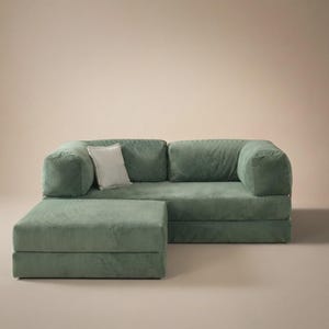 May include: A sage green corduroy sofa with a matching ottoman and a single white square throw pillow. The sofa features a low-profile design with thick, rounded armrests and a textured fabric finish, providing a structured seating arrangement.