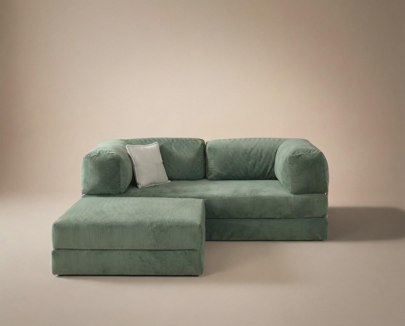 May include: A sage green corduroy sofa with a matching ottoman and a single white square throw pillow. The sofa features a low-profile design with thick, rounded armrests and a textured fabric finish, providing a structured seating arrangement.