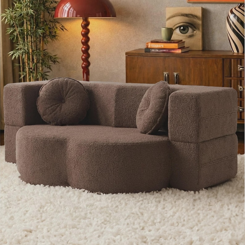 May include: A brown, textured, boucle-style sofa with a curved, clover-shaped seat and a straight backrest. The sofa includes two matching round, tufted throw pillows placed against the backrest, providing a soft, uniform appearance.
