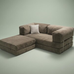 May include: A brown modular sofa with a low-profile design, featuring a rectangular ottoman section, thick cushioned seating, and a single light-colored square throw pillow. The sofa is supported by a thin, metallic frame visible on the side.